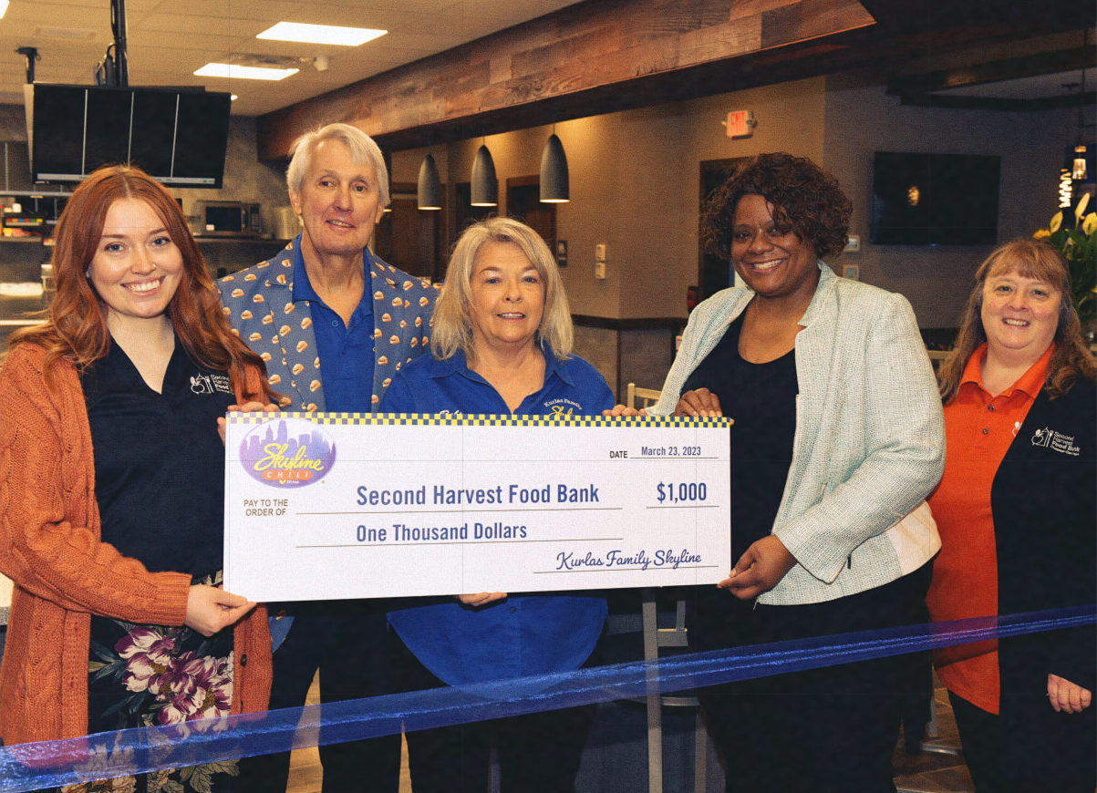 People smiling, standing inside of a Skyline restaurant holding a donation for $1,000