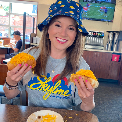 Woman wearing skyline shirt and skyline bucket hat holding coney in each hand